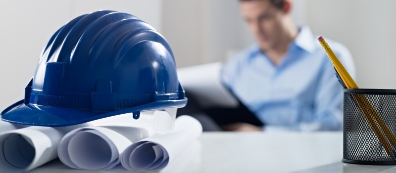 Hardhat and blueprint on desk, with architect in background