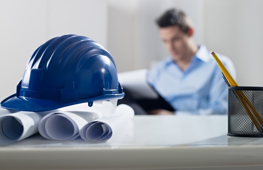 Hardhat and blueprint on desk, with architect in background